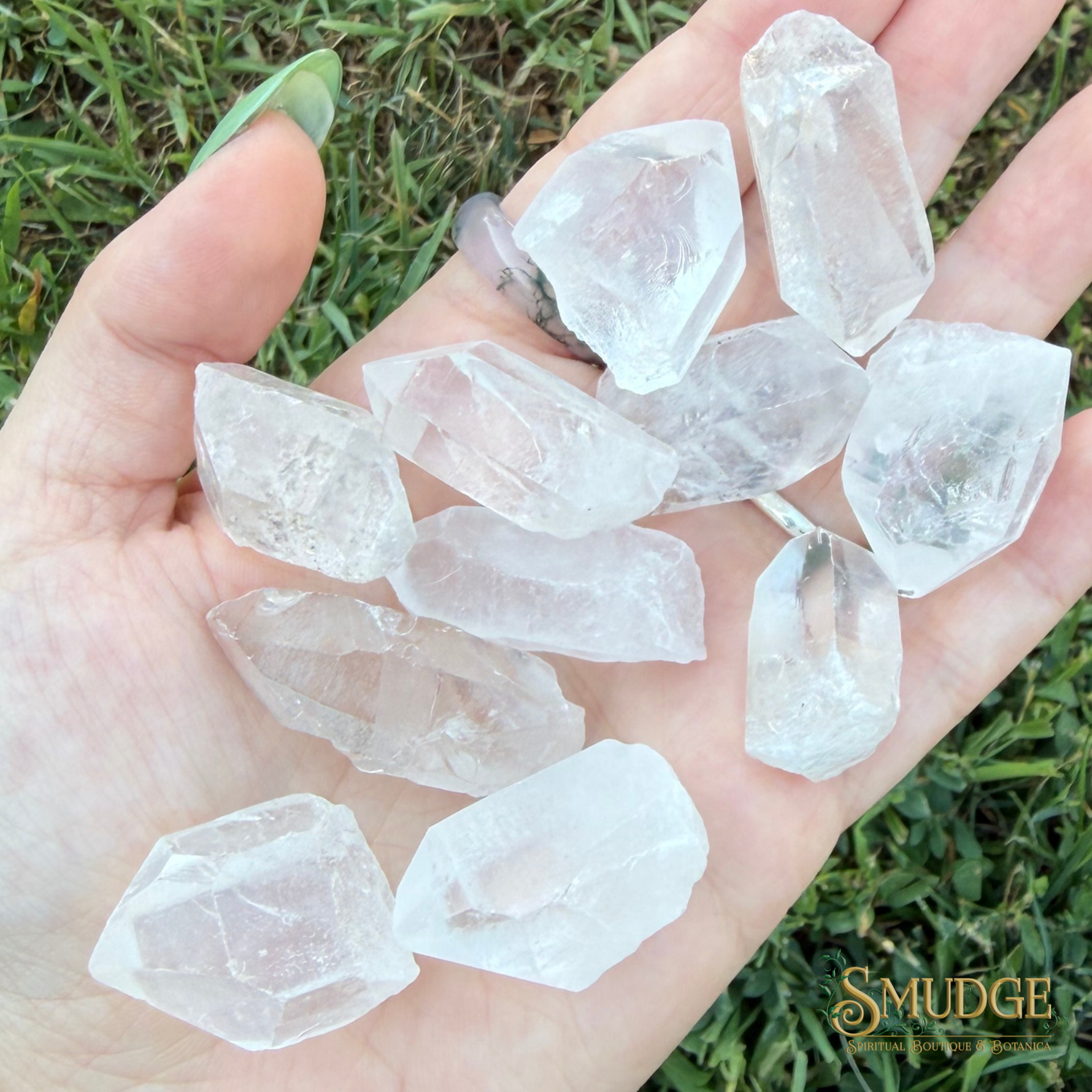 A hand holding multiple raw, rough translucent crystal points over a green grass background. The crystals are clear and semi-translucent.