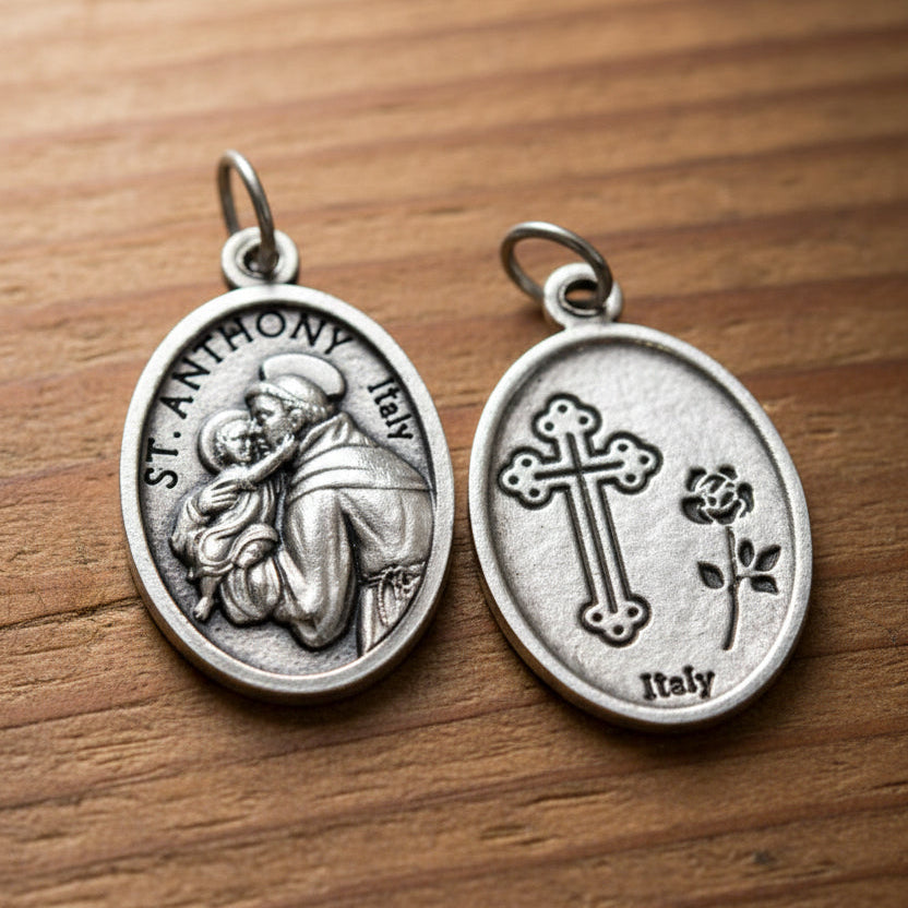 Two silver oval Saint Anthony medal charms shown on a light gray background, with one charm displaying an embossed figure and the other showing a cross and rose motif.