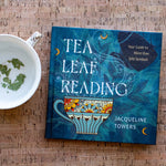 A book titled 'TEA LEAF READING' by Jacqueline Towers lies on a cork surface next to a white teacup with loose tea leaves.