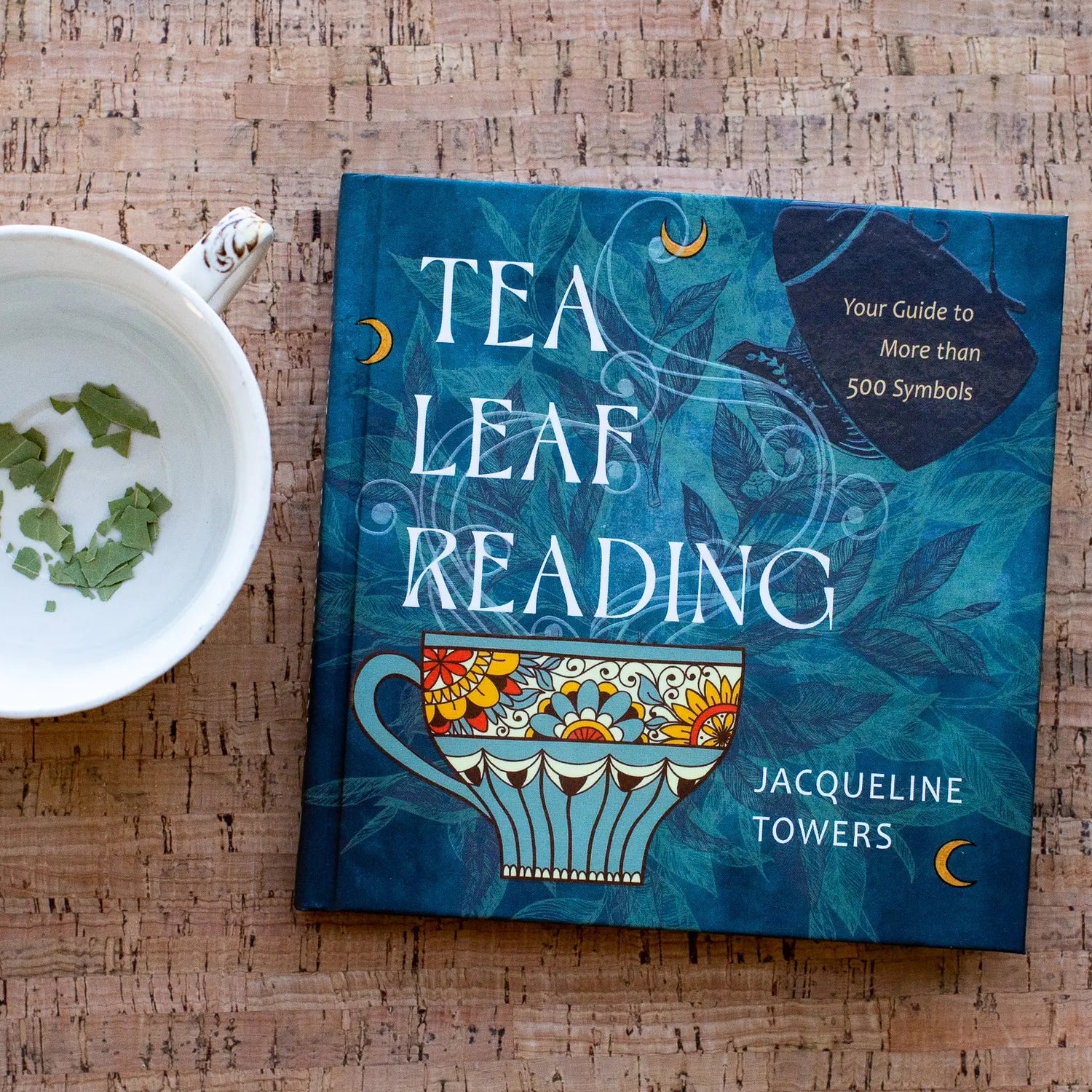 A book titled 'TEA LEAF READING' by Jacqueline Towers lies on a cork surface next to a white teacup with loose tea leaves.