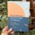 A hand holds a book titled 'The 5-Minute Gratitude Journal' in front of green plants. The book cover is split horizontally with a peach-colored top half and a dark blue-gray bottom half, with white and light blue text.