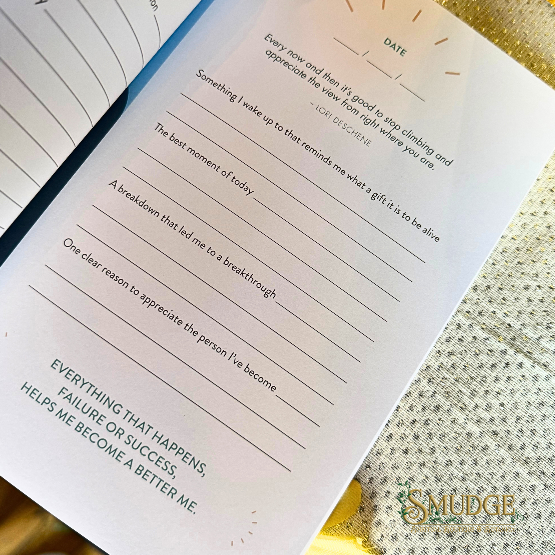 An open journal or notebook showing a reflective writing page with prompts, handwritten text, and the brand name 'SMUDGE' printed in the lower right corner. The page is photographed on a textured gold surface.