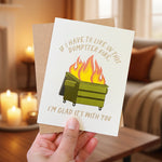 Card with a graphic of a dumpster on fire and text, held by a hand against a white background.