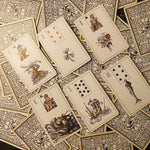 A spread of vintage playing cards with ornate black and white illustrated backs and face cards showing detailed historical scenes on a dark textured surface.