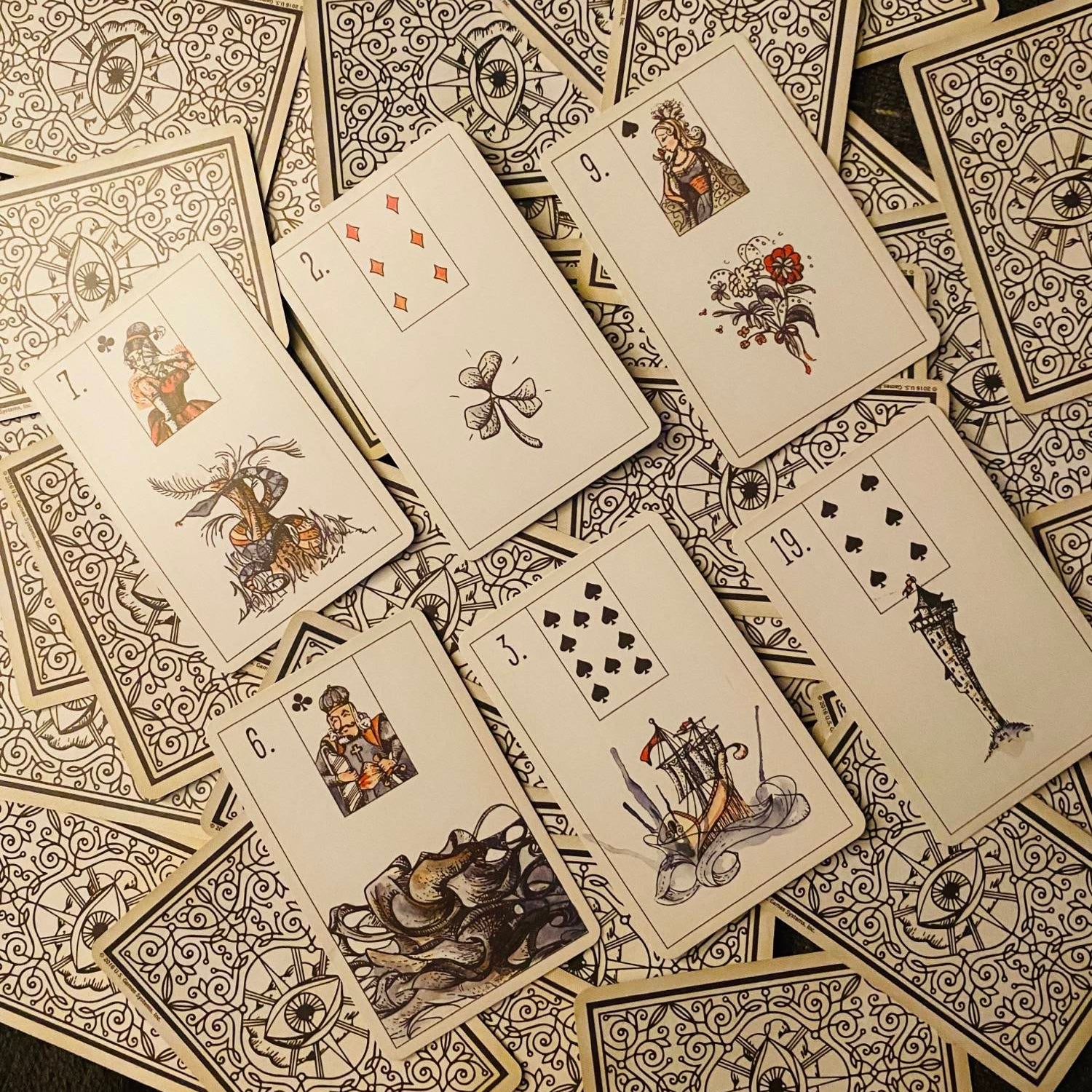 A spread of vintage playing cards with ornate black and white illustrated backs and face cards showing detailed historical scenes on a dark textured surface.