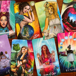 A set of illustrated tarot or oracle cards spread out on a wooden surface, showing colorful fantasy-style artwork with women, animals, celestial elements, and text such as 'Queen of Cups' and '8 of Pentacles.'.