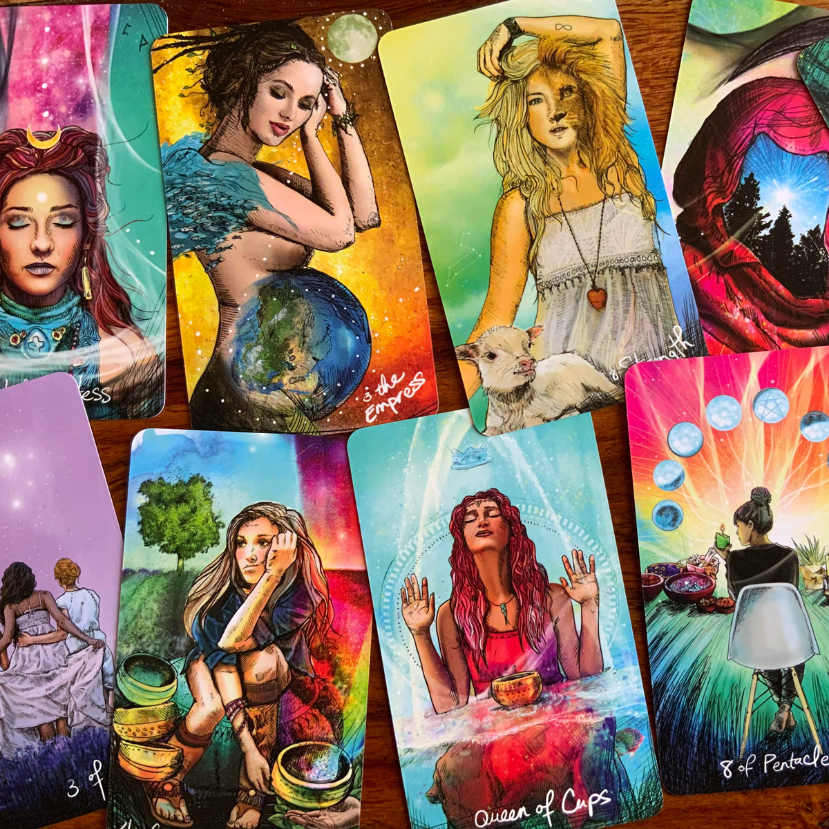 A set of illustrated tarot or oracle cards spread out on a wooden surface, showing colorful fantasy-style artwork with women, animals, celestial elements, and text such as 'Queen of Cups' and '8 of Pentacles.'.
