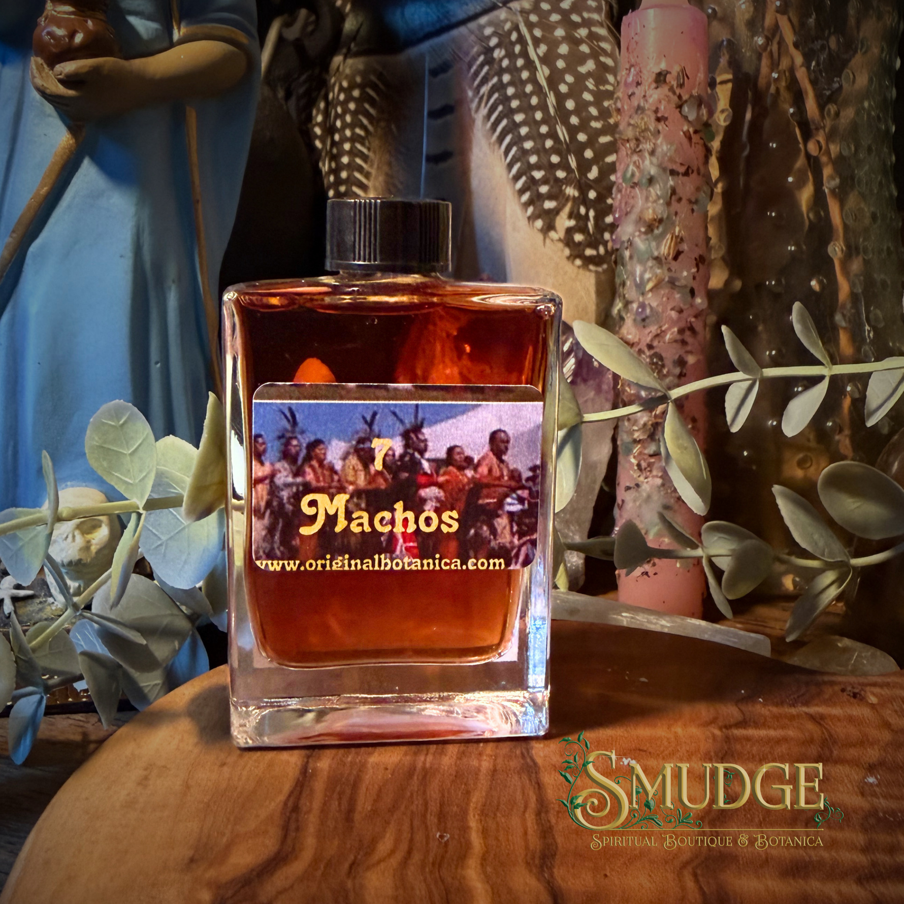 A square glass bottle of Machos spiritual fragrance oil by Original Botany, filled with amber liquid and topped with a black cap, displayed on a wooden surface with decorative botanical and figurine props.