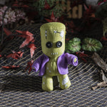Green plush toy with a purple cape on a textured surface with dried leaves and flowers.