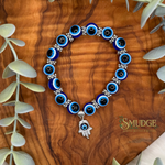 A beaded bracelet with blue and white evil eye beads, silver spacer details, and a silver Hamsa charm, displayed on a wooden surface with green eucalyptus leaves. The brand logo 'SMUDGE' is visible in the bottom right corner.