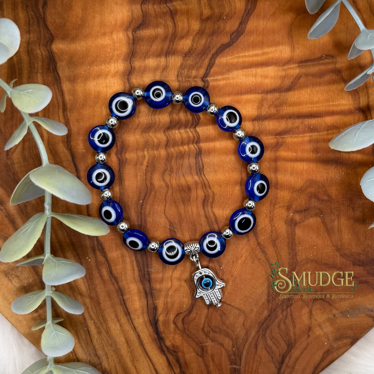 A beaded stretch bracelet with blue, white, and black evil eye beads and a silver Hamsa charm, displayed on a wooden board with eucalyptus leaves. The brand logo 'SMUDGE' is visible in the bottom right corner.