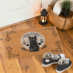 A rectangular coir doormat with a black cat, crescent moons, stars, and the word 'WELCOME' in the center, styled on a wooden floor near a door with a lantern, woven basket, towel, plant, and black sneakers.