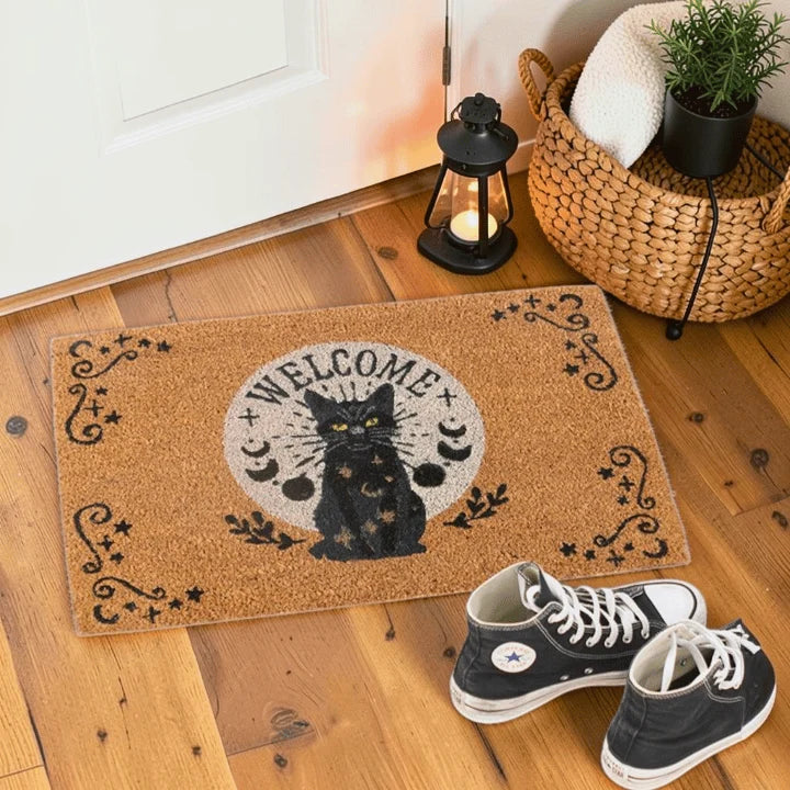 A rectangular coir doormat with a black cat, crescent moons, stars, and the word 'WELCOME' in the center, styled on a wooden floor near a door with a lantern, woven basket, towel, plant, and black sneakers.