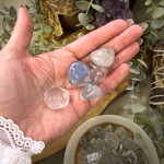 A hand holds several polished tumbled stones, including clear quartz and a blue stone, with more stones in a bowl and crystals in the background among greenery.