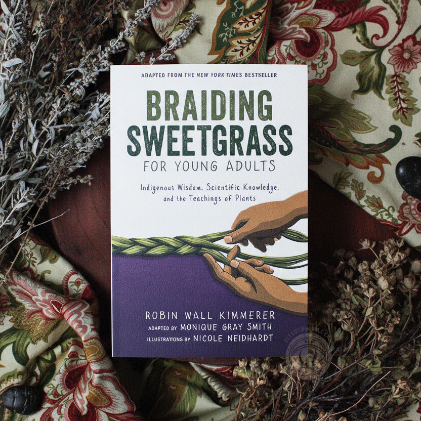 The book 'Braiding Sweetgrass for Young Adults' by Robin Wall Kimmerer is displayed flat on a patterned textile background. The cover features a white top half with dark green text and a purple bottom half with an illustration of hands holding a braided green plant.
