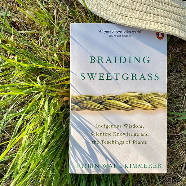 The book 'Braiding Sweetgrass' by Robin Wall Kimmerer lies on a bed of green grass, with a woven straw hat partially visible at the top right. The book cover features a white background with green and gold text and an illustration of a braided grass bundle.