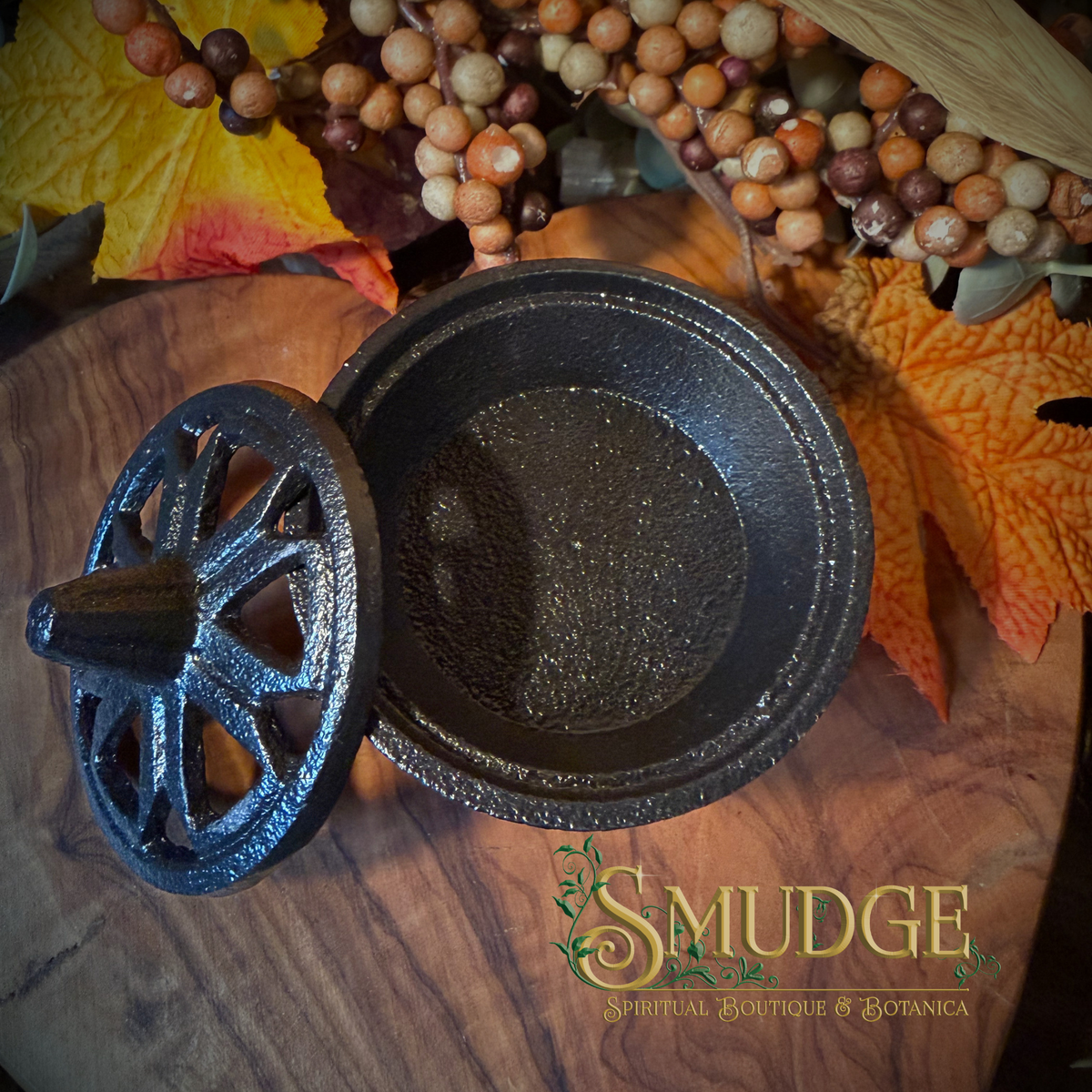 A dark cast iron cauldron-shaped pot with its matching lid leaning beside it on a wooden surface, surrounded by autumn leaves, berries, and a visible 'SMUDGE Spiritual Boutique & Botanica' logo watermark.