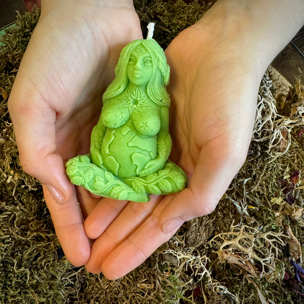 A green sculpted candle shaped like a seated female figure with carved floral and celestial details, held in two hands over a bed of dried moss and botanicals.