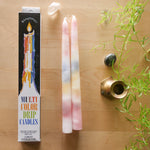 A boxed set of Beautifully Multi Color Drip Candles shown on a light wood surface, alongside two tall pastel wax candles with pink, blue, yellow, and white marbling. A small plant sprig, a polished stone, and a brass holder are placed around the arrangement.