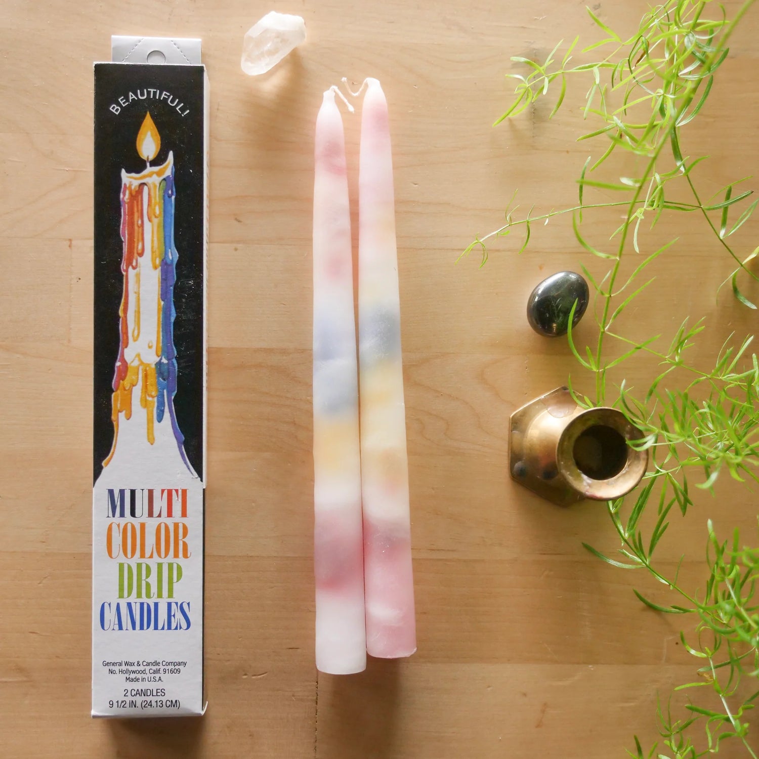 A boxed set of Beautifully Multi Color Drip Candles shown on a light wood surface, alongside two tall pastel wax candles with pink, blue, yellow, and white marbling. A small plant sprig, a polished stone, and a brass holder are placed around the arrangement.