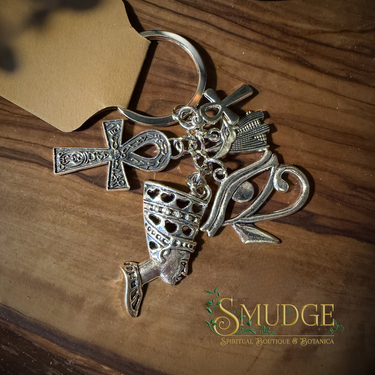 A silver-toned metal keychain featuring multiple ornate charms including an ankh, a stylized eye, a lotus flower, and a small bird, displayed on a wooden surface with a kraft paper corner and the Smudge brand logo visible.