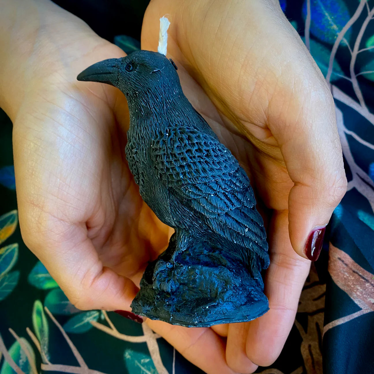A person holds a small black raven figurine in both hands. The figurine has a textured, woven-like surface and stands on a rock-shaped base.