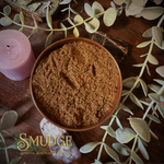 Brown powder incense powder in a bowl surrounded by green leaves and a pink candle on a wooden surface, with 'Smudge' branding.