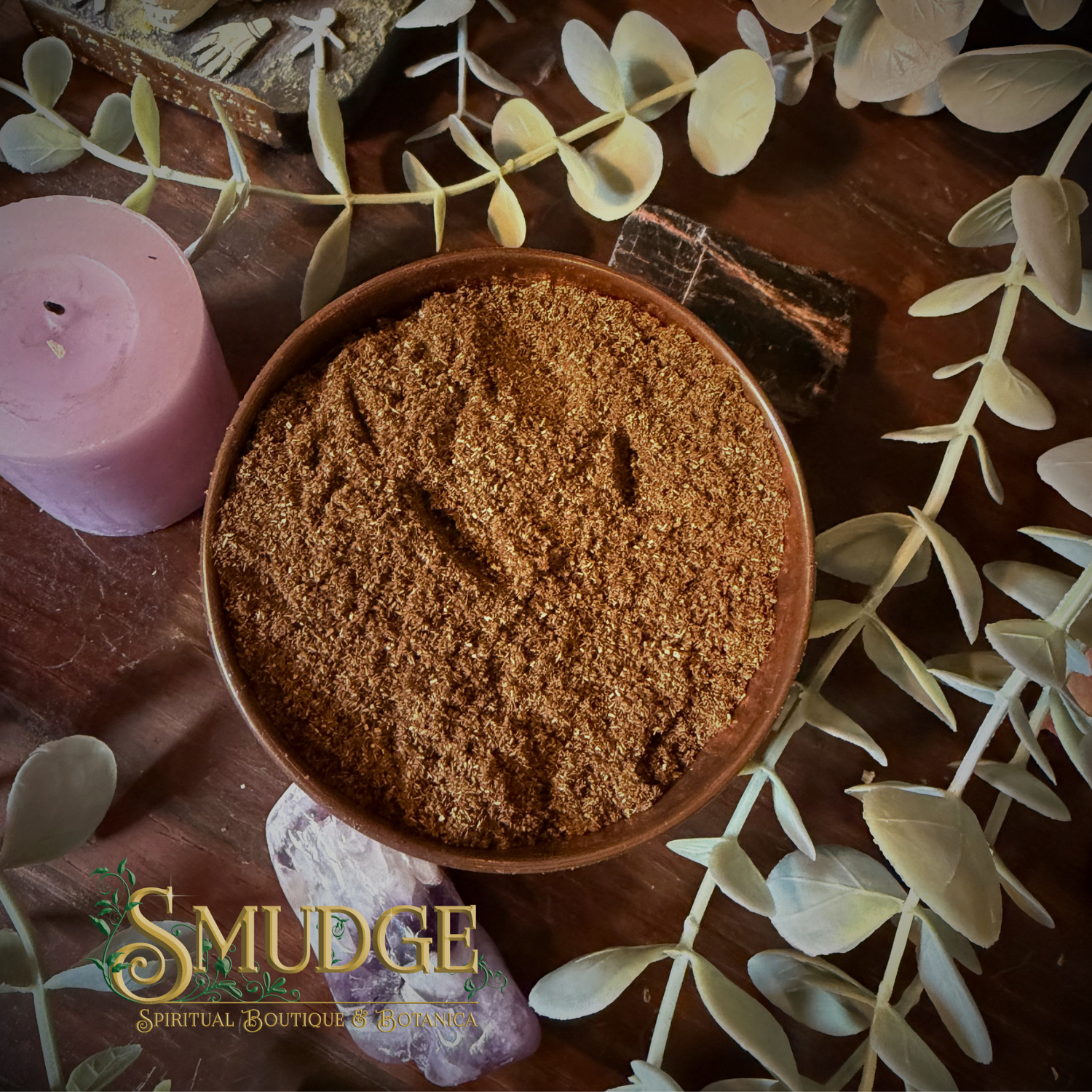 Brown powder incense powder in a bowl surrounded by green leaves and a pink candle on a wooden surface, with 'Smudge' branding.