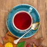 A top-down view of a blue speckled ceramic cup and saucer filled with red tea, styled with a gold spoon, autumn leaves, berries, and a small yellow fruit on a blue wooden surface.