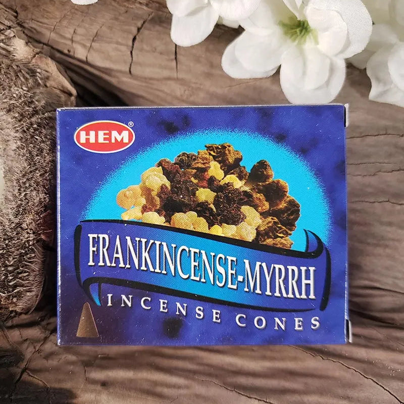 A box of HEM Frankincense Myrrh Incense Cones placed on a rustic wooden surface with white flowers in the background.