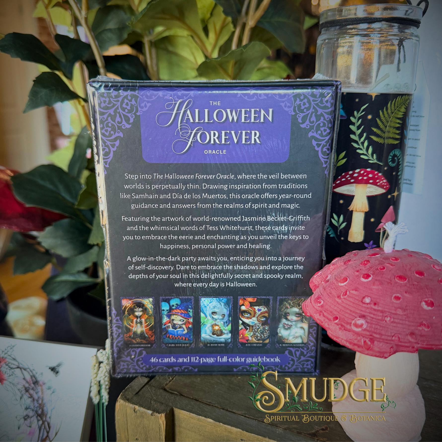 The back cover of 'The Halloween Forever Oracle' card deck box is shown, featuring purple and lavender artwork with descriptive text. The box is displayed on a tabletop next to decorative mushrooms and a glass jar, with a 'SMUDGE Spiritual Boutique & Retreats' logo at the bottom.
