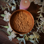 Wooden bowl with brown incense powder on a decorative surface with flowers and a candle, featuring the brand 'Smudge'.