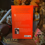 The back cover of a book titled 'Hoodoo Cleansing and Protective Magic' by Miss Aida, featuring a bright orange background with black text and a small photo of a character. The book is displayed on a dark, mystical surface surrounded by autumn-themed decorations, including a gold bust, a pink feather, and a spider web.