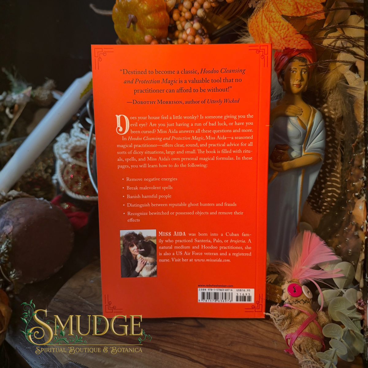 The back cover of a book titled 'Hoodoo Cleansing and Protective Magic' by Miss Aida, featuring a bright orange background with black text and a small photo of a character. The book is displayed on a dark, mystical surface surrounded by autumn-themed decorations, including a gold bust, a pink feather, and a spider web.