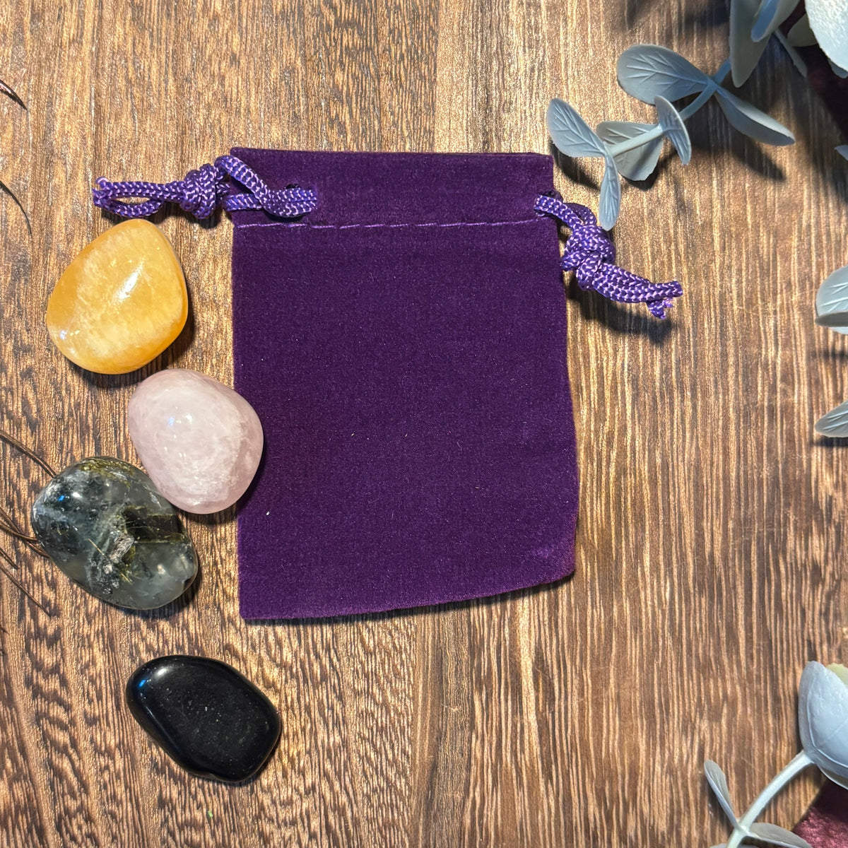Purple velvet drawstring bag on a wooden surface with crystals and plants.