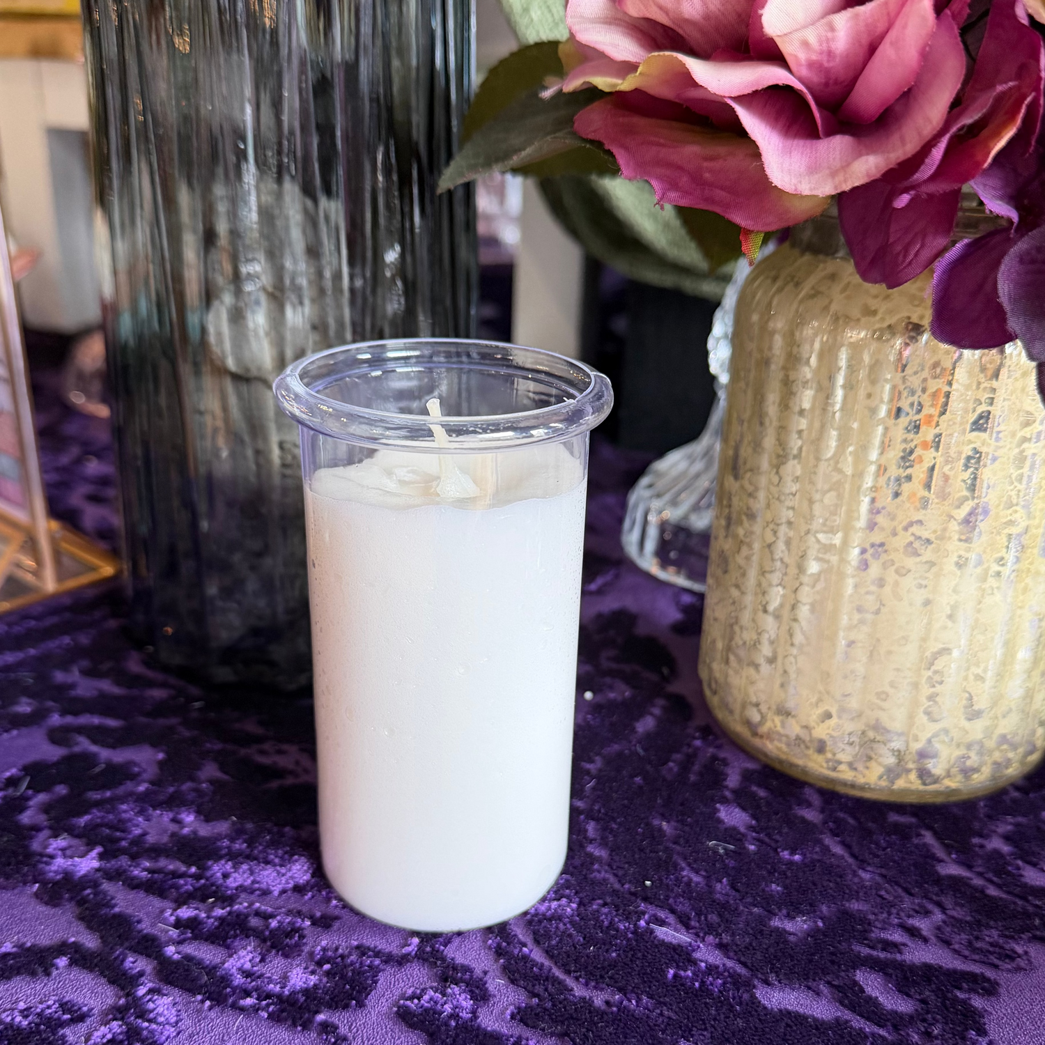 White candle in a clear container on a purple surface with a vase of flowers in the background.