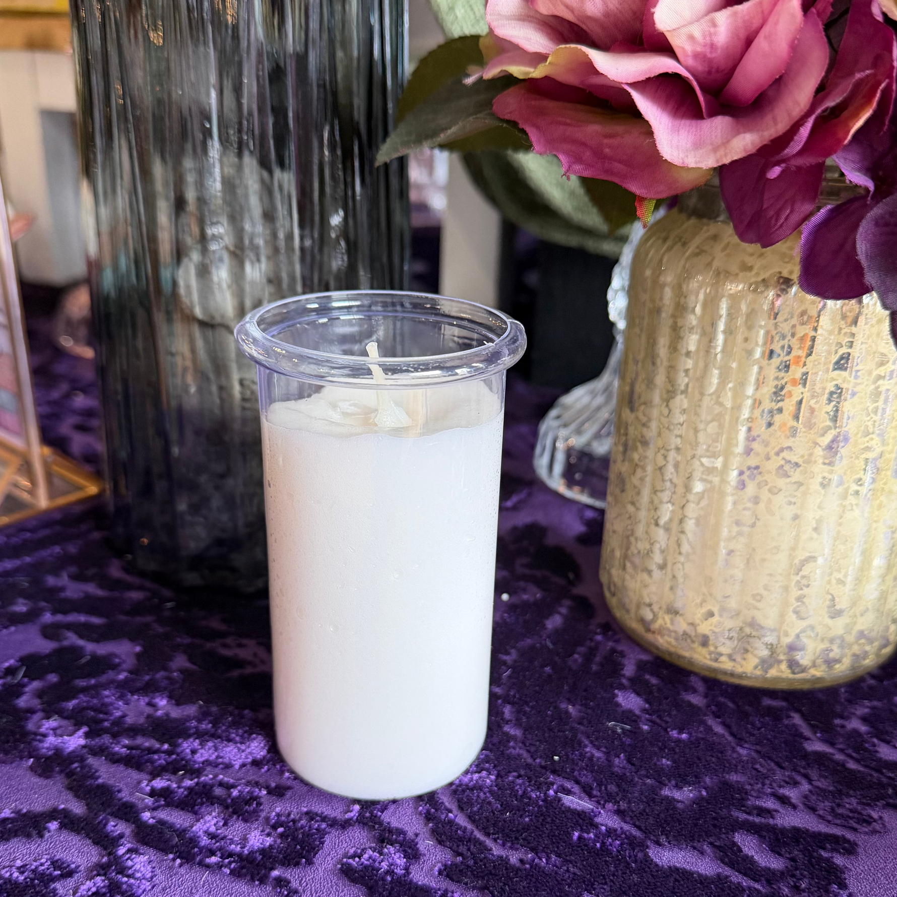 White candle in a clear container on a purple surface with a vase of flowers in the background.