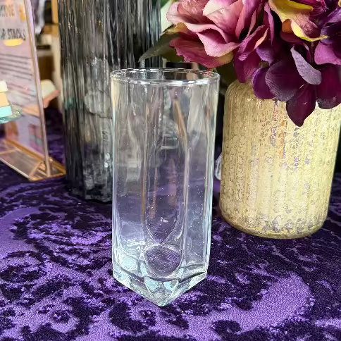 Clear glass on a purple patterned tablecloth with a vase of flowers in the background
