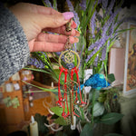 Hand holding a decorative keychain with red beads and a pentacle symbol in front of plants and candles.