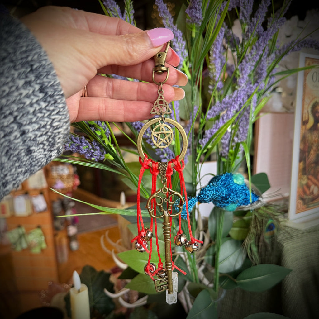 Hand holding a decorative keychain with red beads and a pentacle symbol in front of plants and candles.