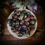 Iridescent stones in a white bowl on a wooden surface with plants and crystals.