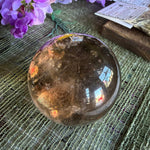 Smoky quartz crystal sphere on a textured surface with purple flowers.