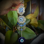 Hand holding a pair of silver and blue evil eye earrings with green leaves in the background
