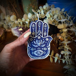 Hand holding a blue Hamsa-shaped candle with decorative elements in the background