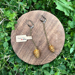 Pine cone-shaped earrings on a wooden stand with a 'Thank You' card, set against a grassy background.