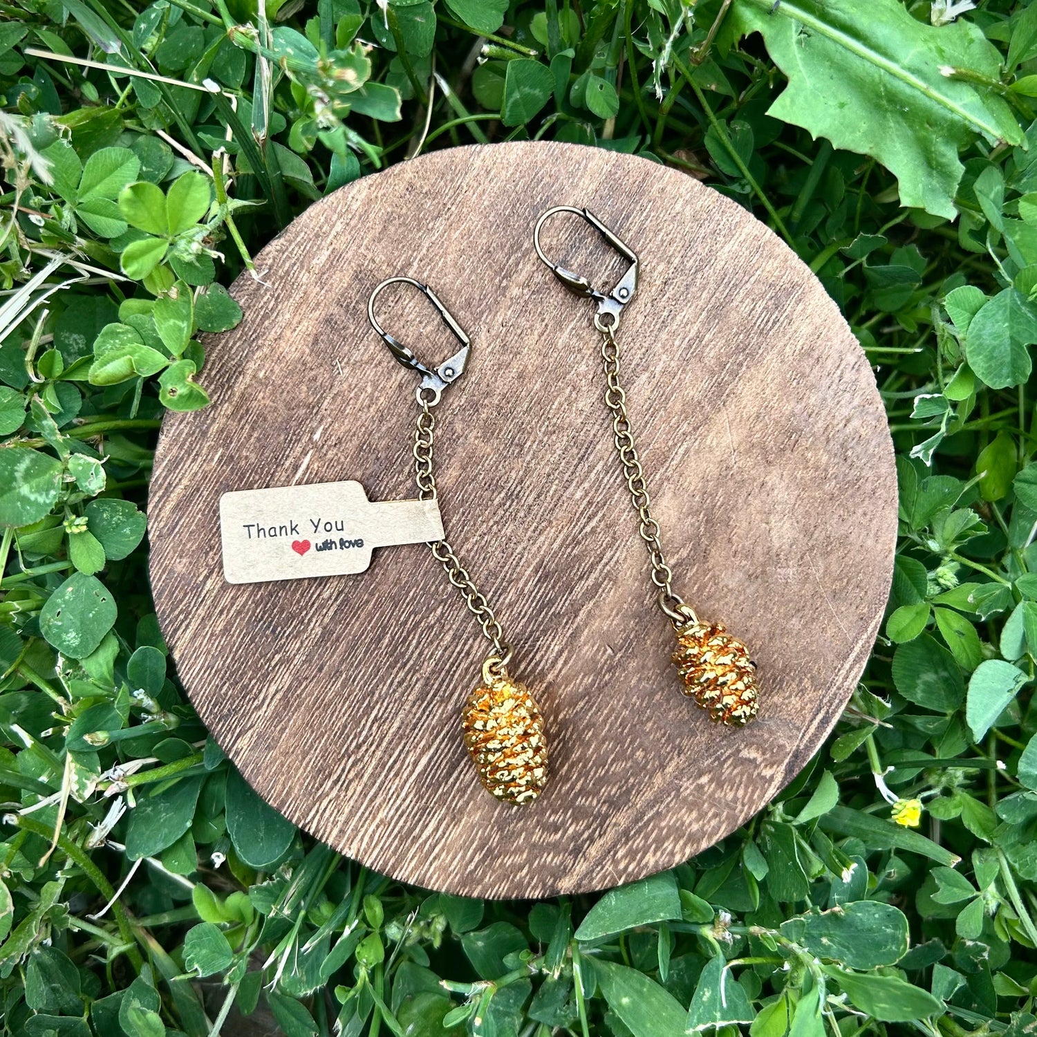 Pine cone-shaped earrings on a wooden stand with a 'Thank You' card, set against a grassy background.