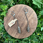 Pine cone-shaped earrings on a wooden round platform with a tag, placed on green grass.