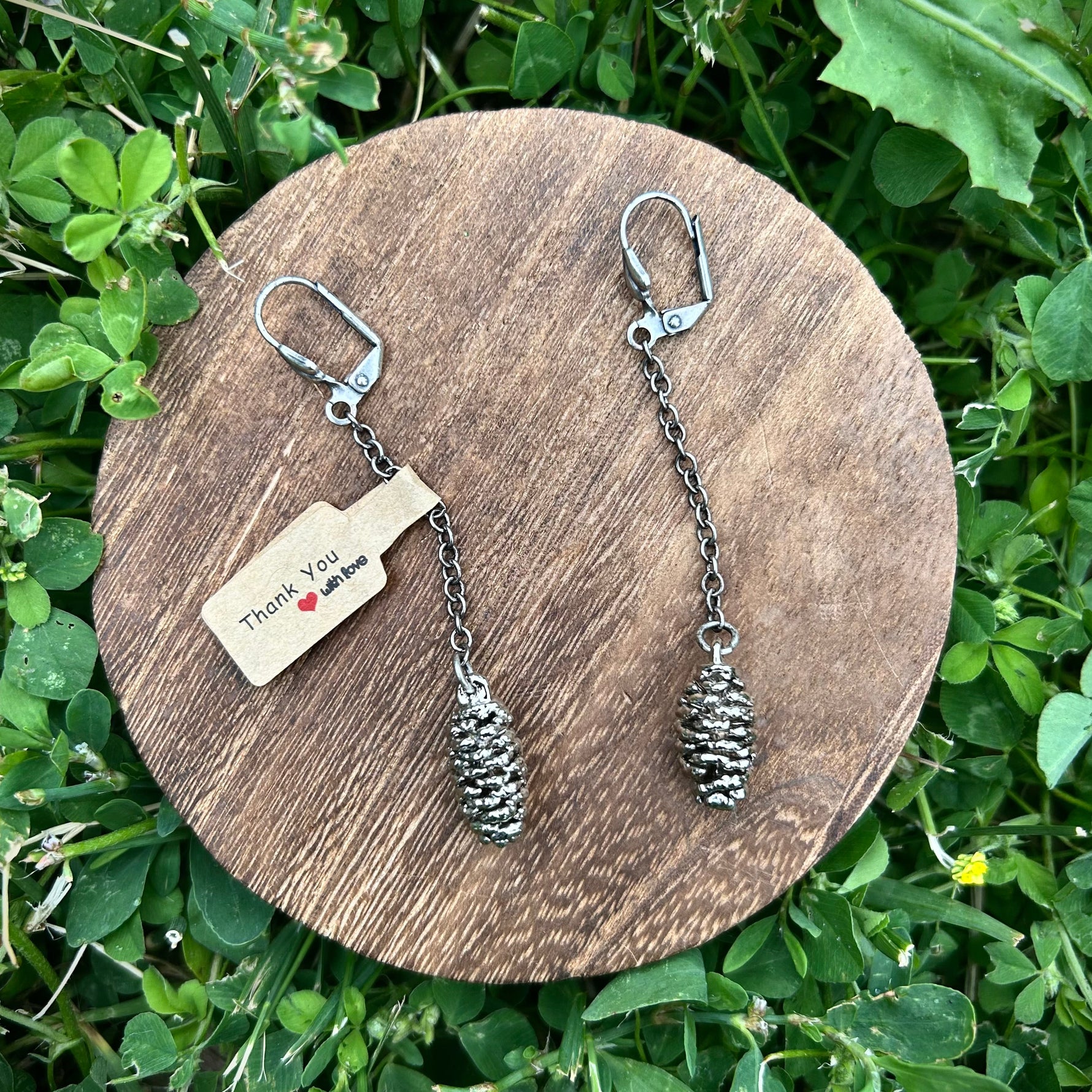 Pine cone-shaped earrings on a wooden round platform with a tag, placed on green grass.