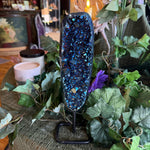 Blue crystal geode on a stand surrounded by greenery and candles in a cozy indoor setting.