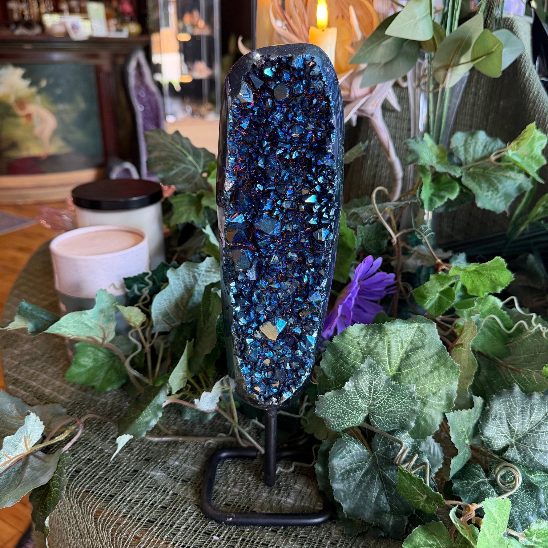 Blue crystal geode on a stand surrounded by greenery and candles in a cozy indoor setting.